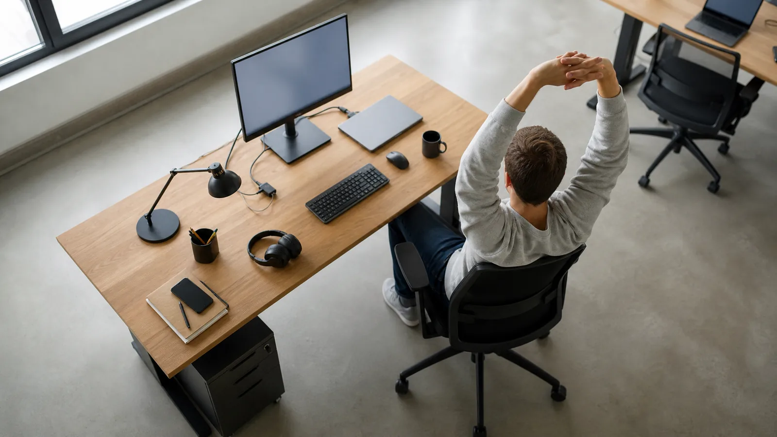 Eine Person sitzt auf einem ergonomischen Bürostuhl am Schreibtisch und dehnt sich, die Arme über dem Kopf verschränkt, von hinten gesehen – Symbolbild zu «Büro-Stretching: 10 Dehnübungen gegen Verspannungen am Schreibtisch»
