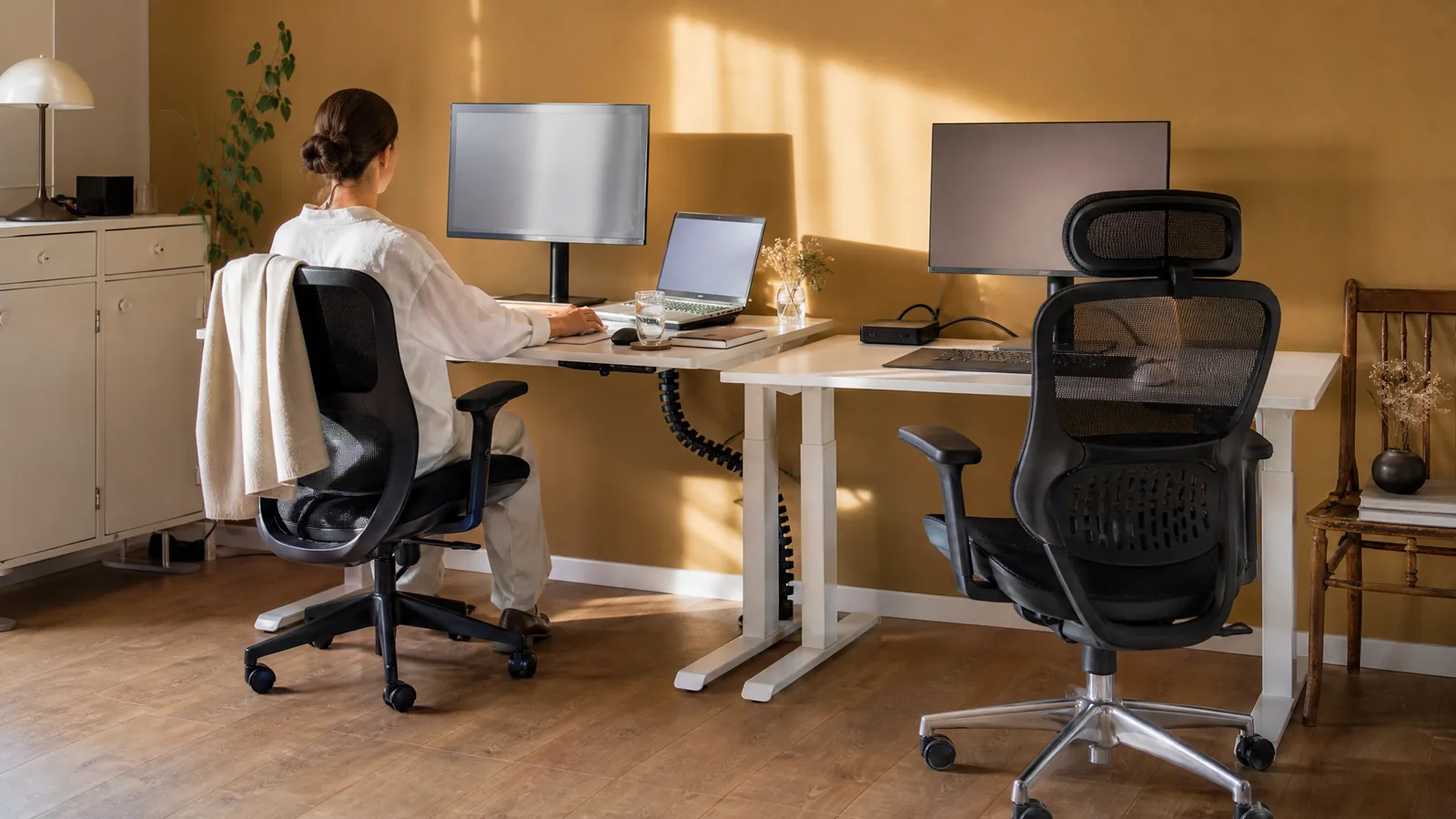 Ein Arbeitsplatz für zwei Personen mit zwei höhenverstellbaren Schreibtischen und ergonomischen Bürostühlen, eine Person arbeitet sitzend, von hinten gesehen – Symbolbild zu «Ergonomischer Arbeitsplatz einrichten: Die komplette Anleitung»
